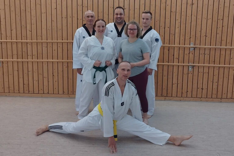 Sechs Personen im Dojo, eine zeigt Spagat, andere tragen Kampfsportanzüge mit Gürteln.