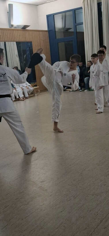 Kind zeigt hohen Kick auf Schlagpolster, andere im weißen Dobok beobachten im Trainingsraum.