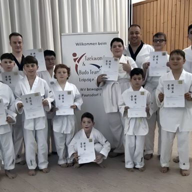 Kinder in weißen Anzügen mit Urkunden, Gruppenfoto vor Taekwondo-&-Budo-Team-Banner.