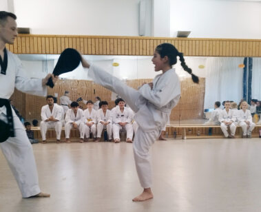 Mädchen zeigt hohen Kick gegen Pratze, Gruppe schaut im Dojang zu.