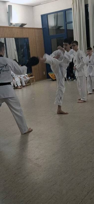 Hoher Kick trifft Schlagpolster, Taekwondo-Training im Dojo, andere beobachten konzentriert.
