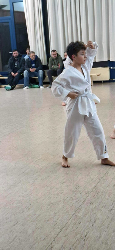 Kind im weißen Anzug zeigt Kampfstellung, Erwachsene beobachten im hellen Trainingsraum.