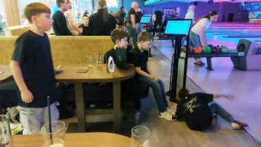 Kind liegt vor Ballrückgabe, andere sitzen am Tisch, bunte Bowlingbahn im Hintergrund.