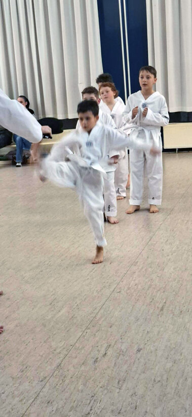 Kind zeigt Frontkick zur Taekwondo-Prüfung. Dahinter warten die restlichen Prüflinge in weißen Anzügen.