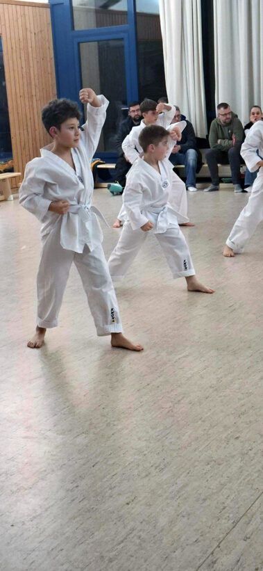 Kinder zeigen synchron Grundschule im Dojo, weiße Anzüge, Zuschauer sitzen im Hintergrund.