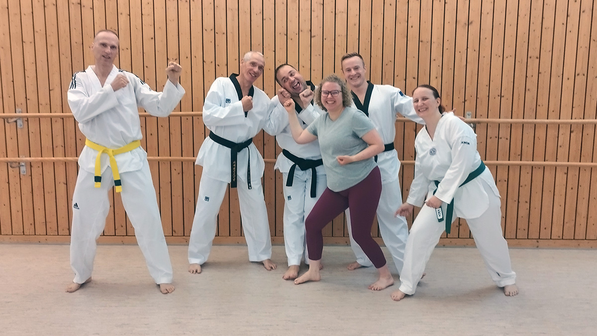 Sechs Personen posieren im Dojo, fünf in Doboks, eine in Sportkleidung, alle lächeln.