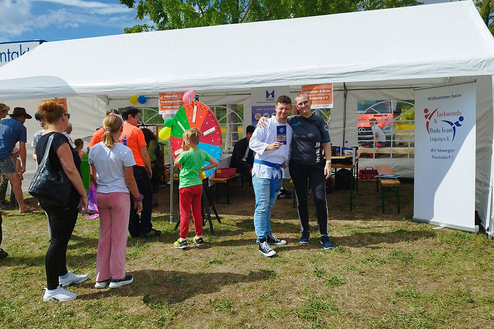 Vereinsstand auf Stadtteilfest mit Besuchern, Glücksrad, Taekwondo-Banner und guter Stimmung.