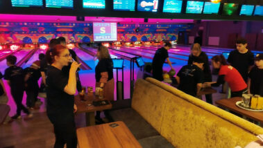 Bowlingbahn mit bunter Beleuchtung, Leute trinken und spielen, „SPLIT“-Anzeige im Hintergrund.