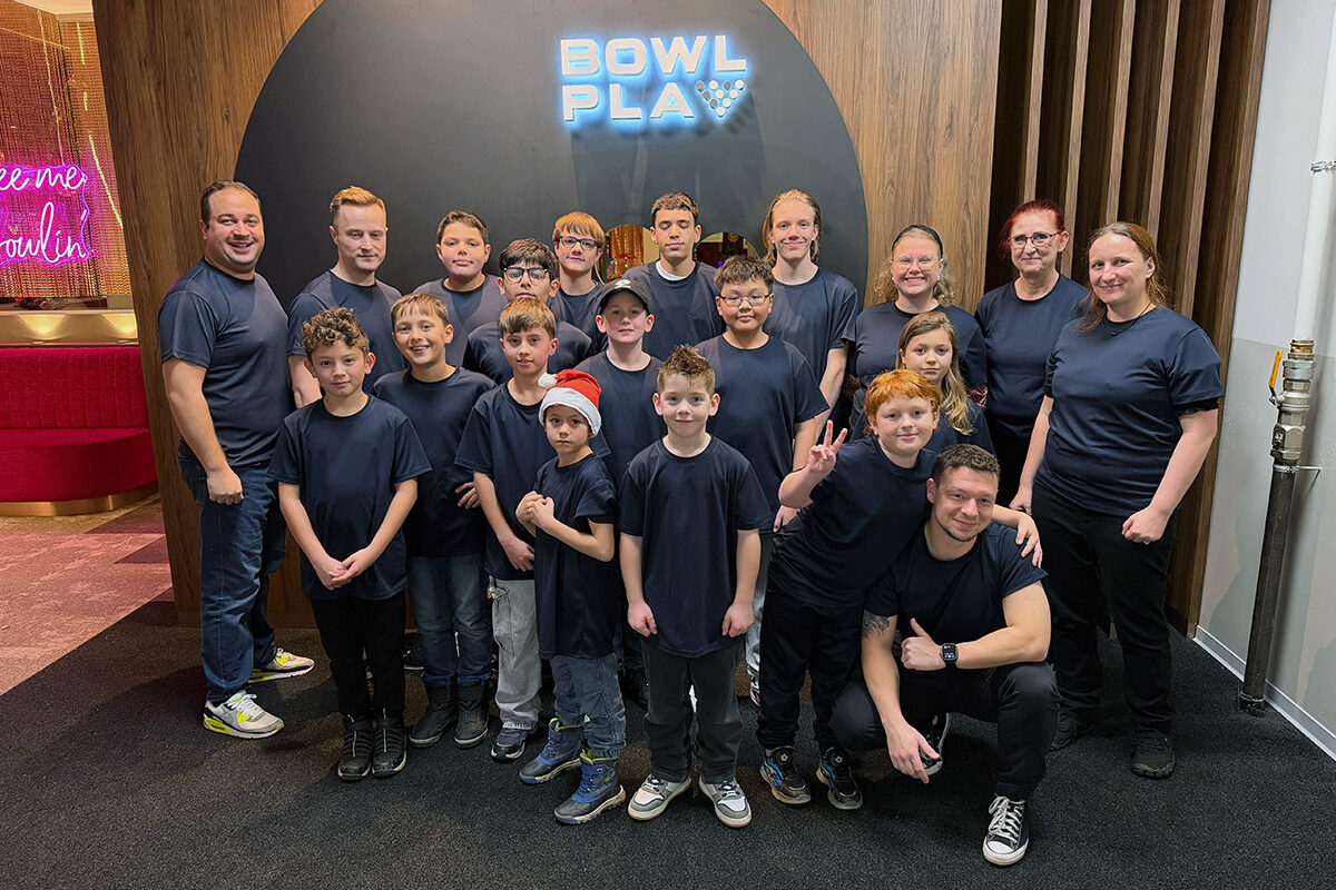 Gruppe in blauen Shirts posiert fröhlich vor „BOWL PLAY“-Leuchtschild, Bowling-Event.