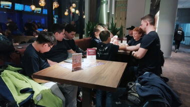 Gruppe sitzt am Tisch im Café, dunkle Shirts, warme Beleuchtung.