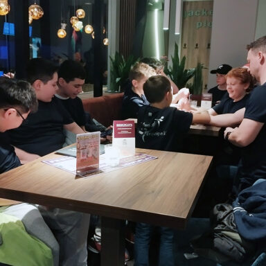 Gruppe sitzt am Tisch im Café, dunkle Shirts, warme Beleuchtung.