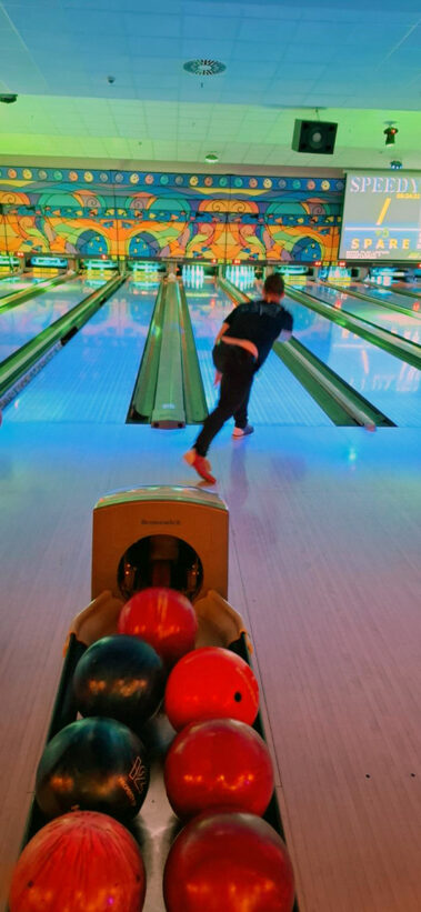 Person beim Bowling, bunte Beleuchtung, Ballrückgabe mit Kugeln, „SPARE“-Anzeige im Hintergrund.