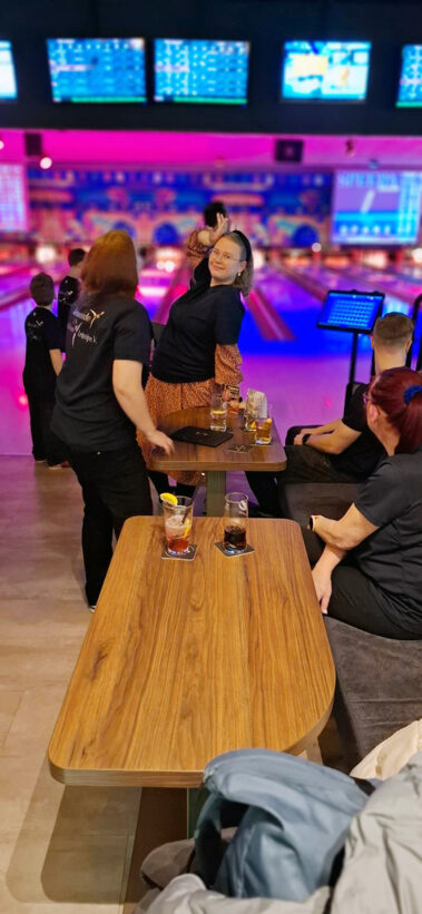 Gruppe beim Bowling, bunte Beleuchtung, Getränke auf Tischen, entspannte Stimmung.