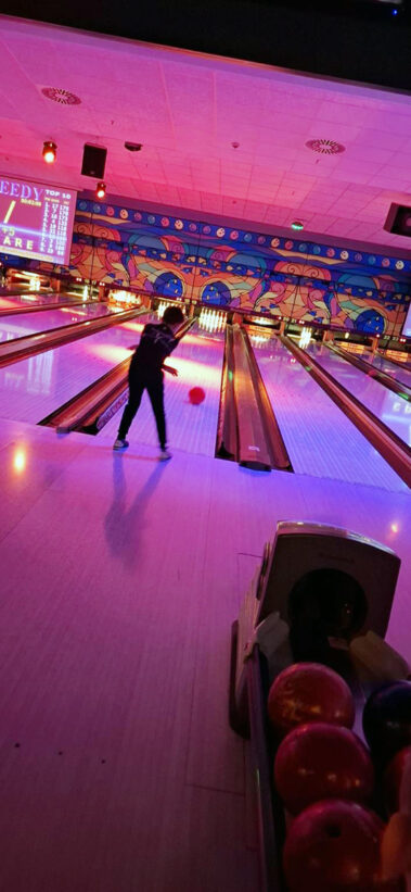 Person wirft rote Bowlingkugel, bunte Beleuchtung, Scoreboard und Ballrückgabe im Vordergrund.