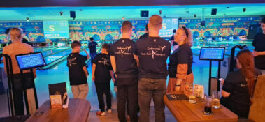 Team in blauen Shirts beim Bowling, bunte Wandkunst und „SPEEDY SPLIT“-Anzeige im Hintergrund.