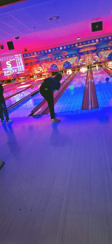 Person wirft Bowlingkugel, Bahn leuchtet blau, bunte Wandkunst und Scoreboard im Hintergrund.