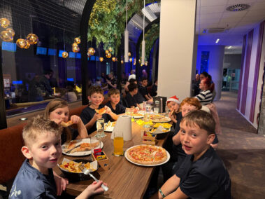Kinder essen Pizza und Pommes am langen Tisch, festliche Stimmung mit Weihnachtsmütze.