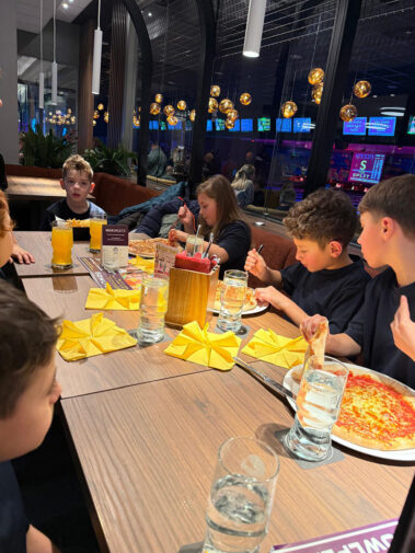 Kinder essen Pizza am großen Tisch, gelbe Servietten gefaltet, Bowlingbahn im Hintergrund.