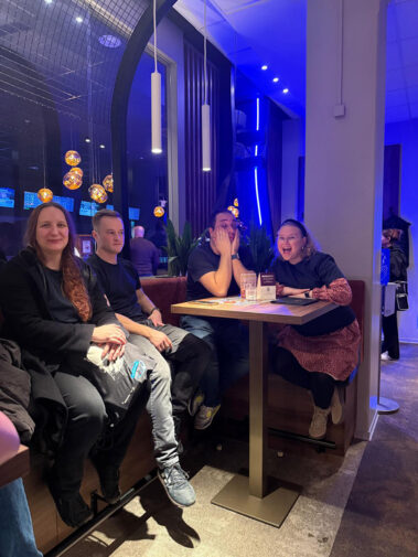 Vier Personen am Tisch im Lokal, entspannte Stimmung, Drinks und Menü auf dem Holztisch.