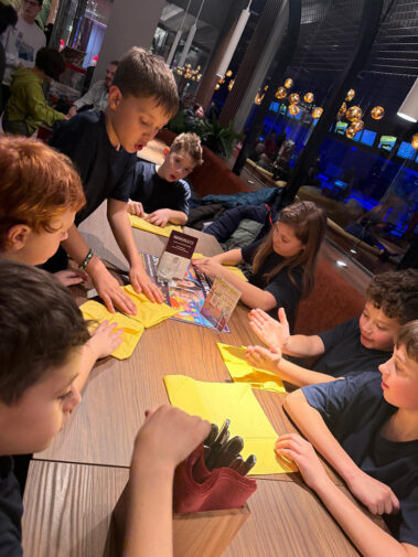 Kinder sitzen am Tisch mit gelben Servietten, warmes Licht, moderne Deko im Hintergrund.