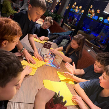 Kinder sitzen am Tisch mit gelben Servietten, warmes Licht, moderne Deko im Hintergrund.