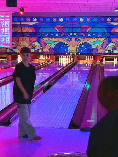 Kind steht vor leuchtender Bowlingbahn, bunte Wandkunst und „TOP 10“-Anzeige im Hintergrund.