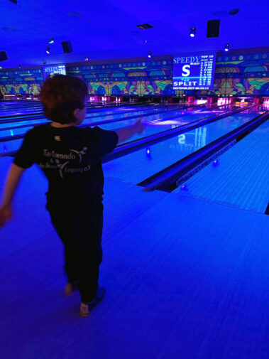 Kind wirft Bowlingkugel, leuchtende Bahn, „SPLIT“-Anzeige und Top-10-Scores im Hintergrund.