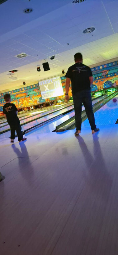 Zwei Personen beim Bowling, bunte Beleuchtung, „STRIKE“ auf dem Bildschirm über der Bahn.