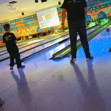 Zwei Personen beim Bowling, bunte Beleuchtung, „STRIKE“ auf dem Bildschirm über der Bahn.
